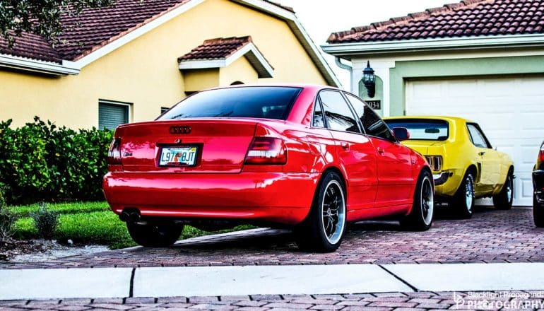 A red and yellow car sit in a driveway outside a yellow house with a green lawn