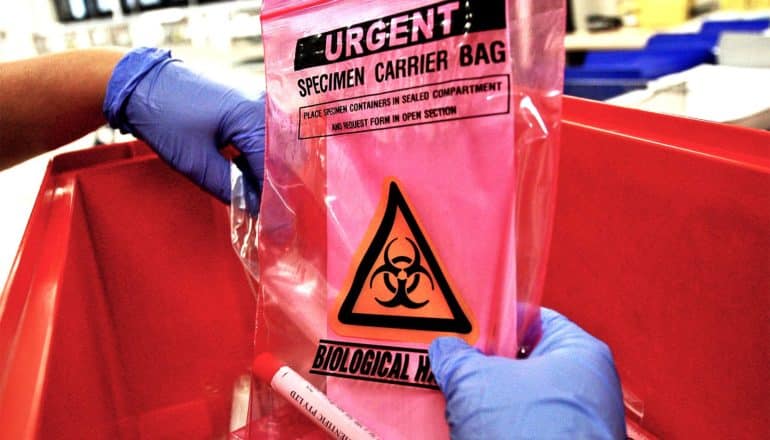 A nurse places a pink specimen carrier bag into a red tub. The bag has the sign for biomedical hazard on it and is labeled "Urgent"