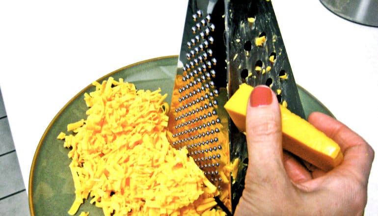 hand holds cheese against metal box grater. Shreds on green plate