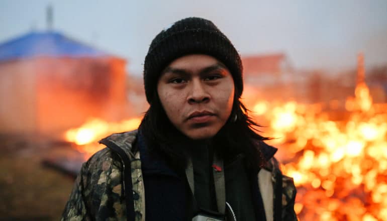 serious man in hat stands in front of burning structures