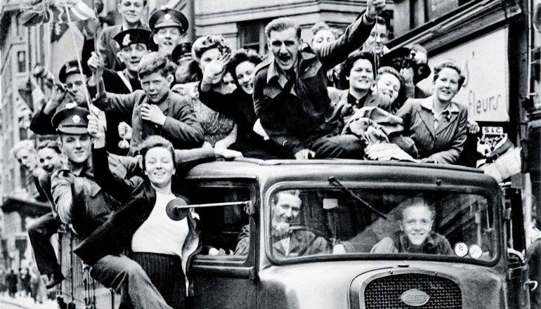 People celebrate while hanging off or standing on a truck rolling down the street on VE Day in 1945