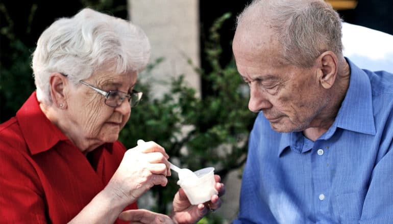 An older woman in red feeds her husband, in blue, from a yogurt cup