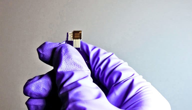 A research wearing a purple glove holds up a tiny black and gold chip