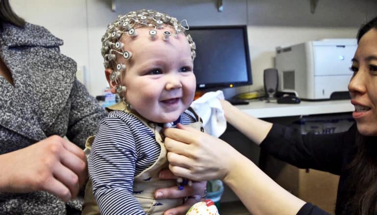 two adults at edge of photo of EEG cap on smiling baby