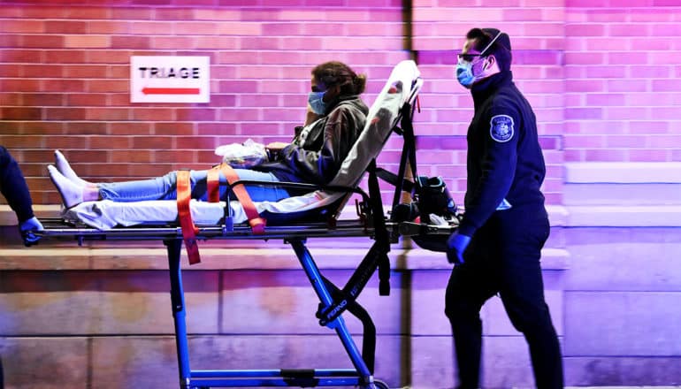 An EMT pushes a young woman on a gurney past a brick wall with a sign that reads "Triage"