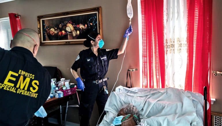 EMTs stand over a man in bed as one hangs an IV bag near a window with bright red curtains