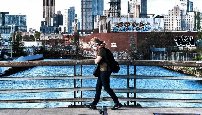 A person in a face mask walks across a bridge with Brooklyn in the background