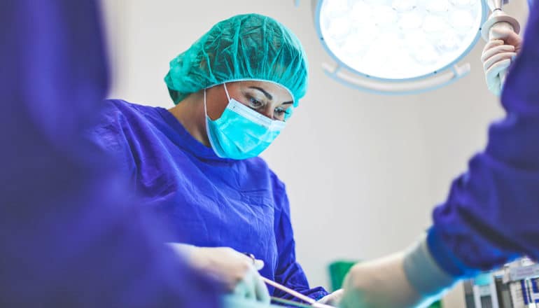 medical professional in cap, mask, scrubs holds forceps under surgical lamp