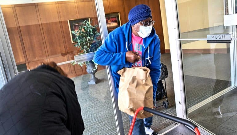 A woman in blue and wearing a medical mask carries a paper back of food through a doorway