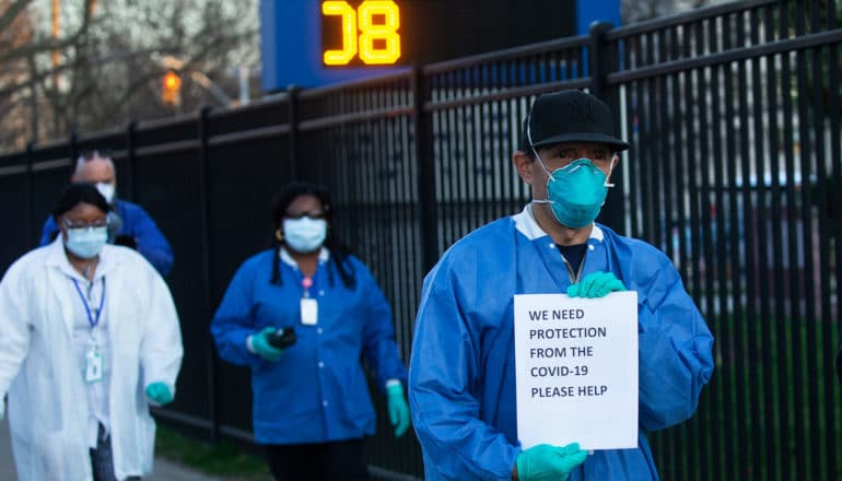 four people in masks and lab coats. Person in front holds sign reading "we need protection from the COVID-19 please help"