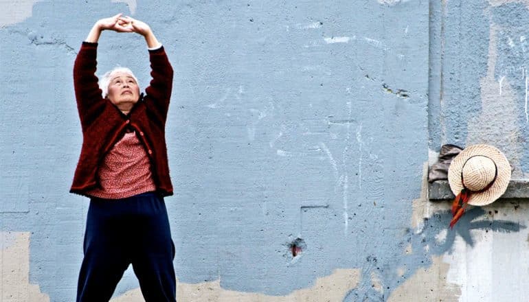 An older woman in red stretches during her exercise while standing against a wall painted blue