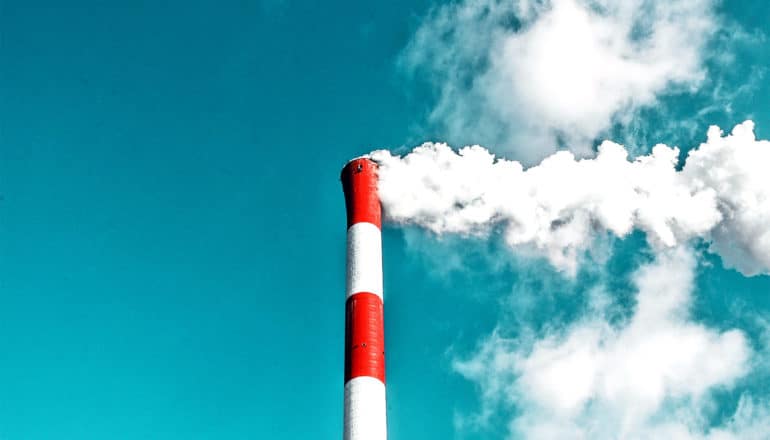 A red and white smokestack spews white smoke into a blue sky