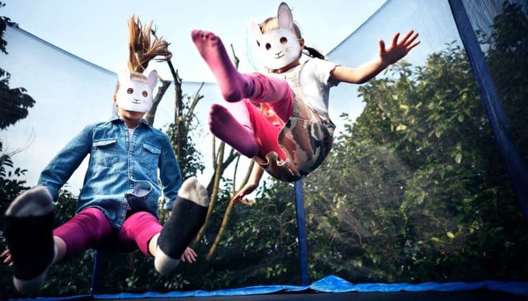 Two young girls in paper rabbit masks jump on a trampoline outside