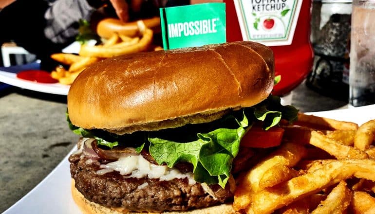 An Impossible burger sits on a plate with fries and a small green flag in the bun that reads "Impossible"