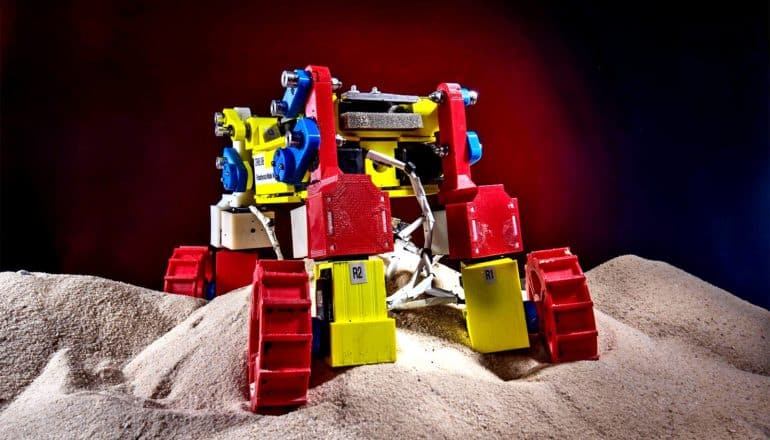 The mini rover stands on sand. It's red, yellow, and blue plastic.