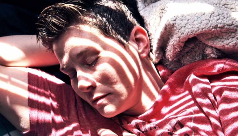 A woman in a red t-shirt sleeps with light coming through blinds onto her face