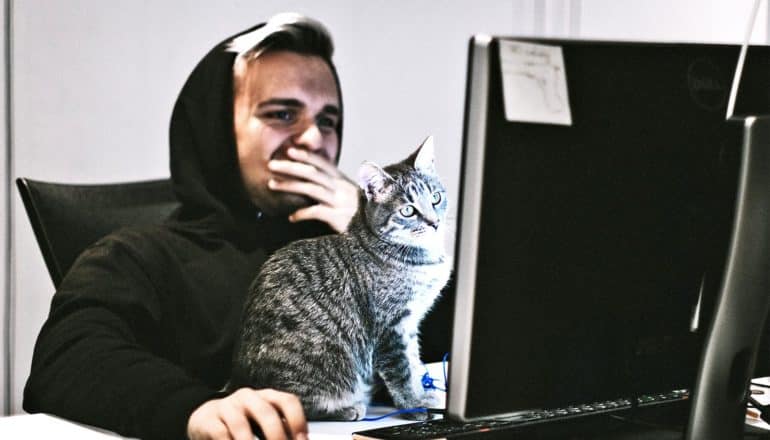 A young man puts his hand over his face as he looks at his computer, along with his cat