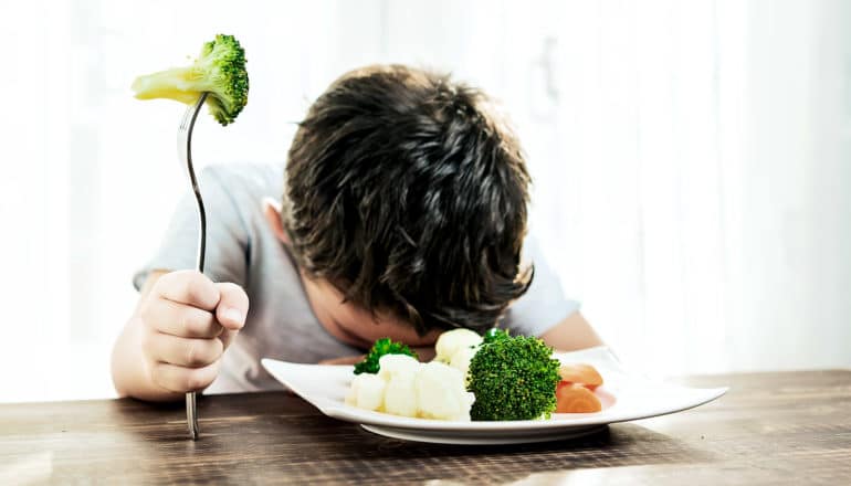 A kid puts his head on the dinner table while holding one piece of broccoli on his fork as a plate of vegetables sits in front of him