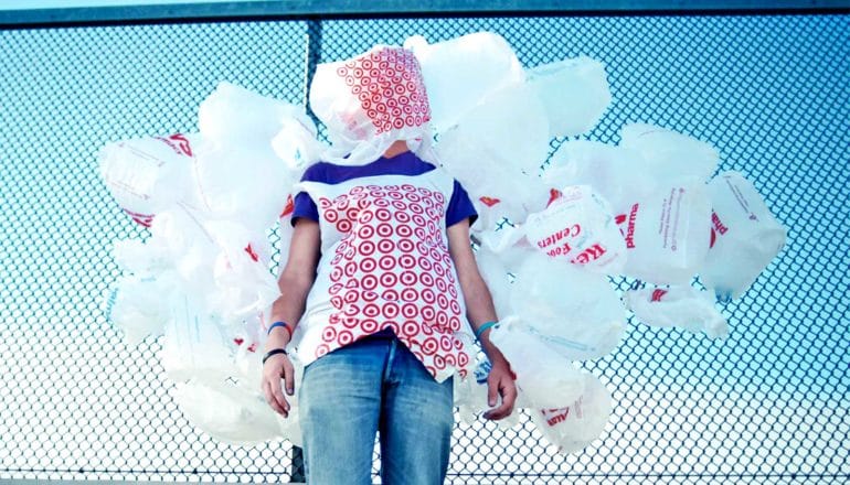 A person stands against a fence, covered in plastic bags