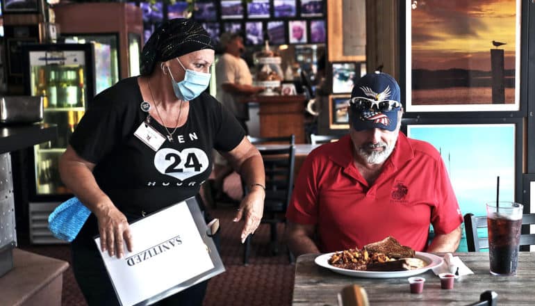 A server brings a customer in a red shirt and ball cap his food while wearing a mask. He looks down at his food.