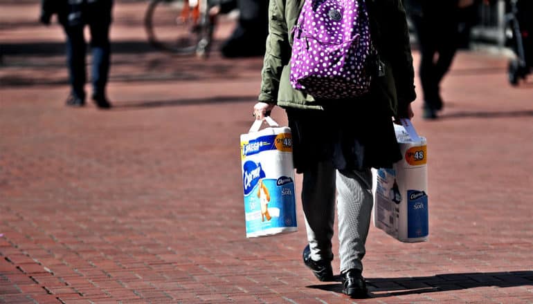 A woman walks on a brick sidewalk with two packages of toilet paper