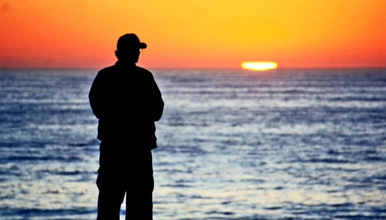 A man stands near the ocean looking out at a sunset