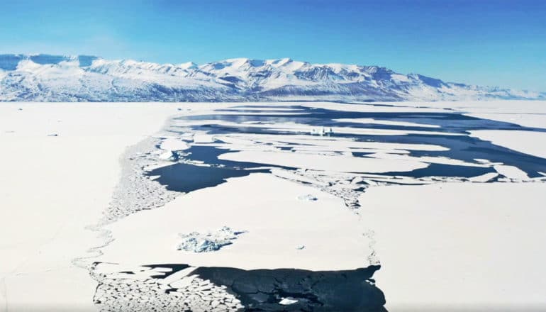 White shorefast ice breaks up to reveal water. There's a mountain range and bright blue sky in the background