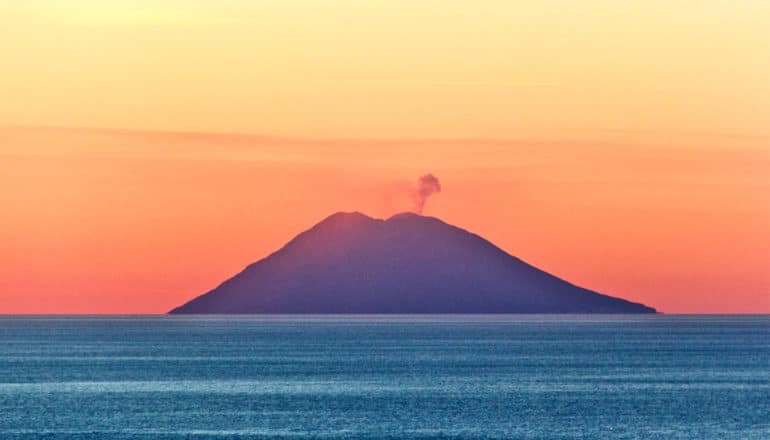 An island volcano has a small plume of smoke coming from its top, with the ocean around it and a reddish orange sunset lighting up the sky