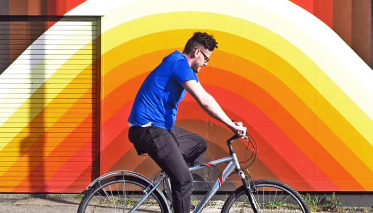A man rides a bike past a wall painted with a color gradient going from brown to red to orange to yellow to white