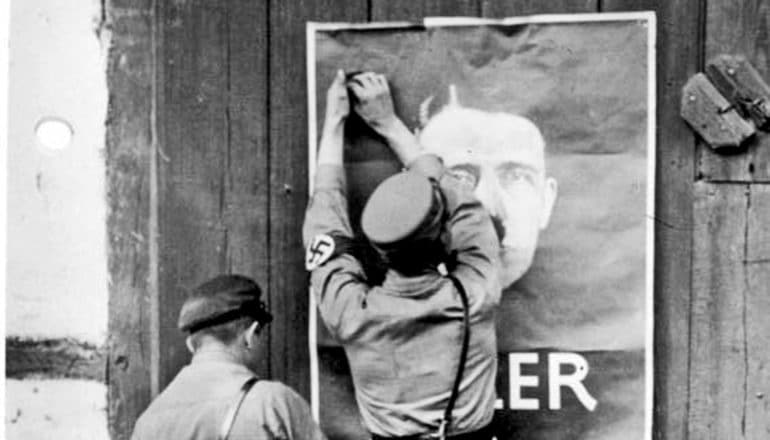 two people, one with swastika armband, hang poster of hitler