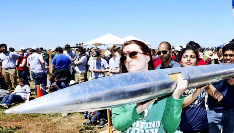 A young woman carries a silver rocket over her shoulder past a crowd