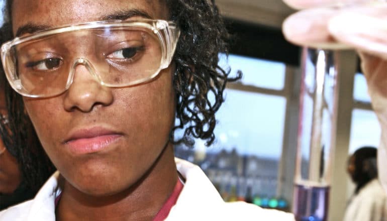 A young woman in a lab coat and glasses looks over at a test tube in her hand