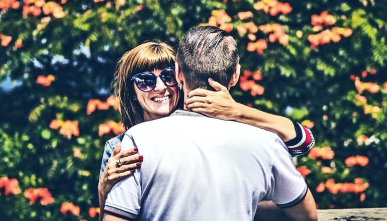 A woman hugs a man while smiling, wearing sunglasses. Orange flowers on green bushes bloom in the background