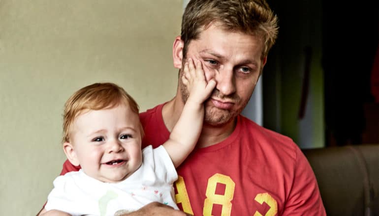 A man holds a baby who's putting his hand on the man's face as he stares off into space with a sad expression