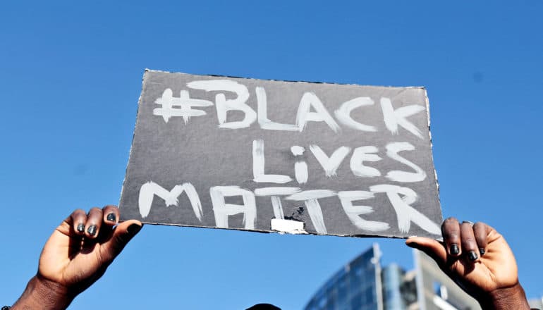 A person holds up a sign that reads "Black Lives Matter" against a bright blue sky