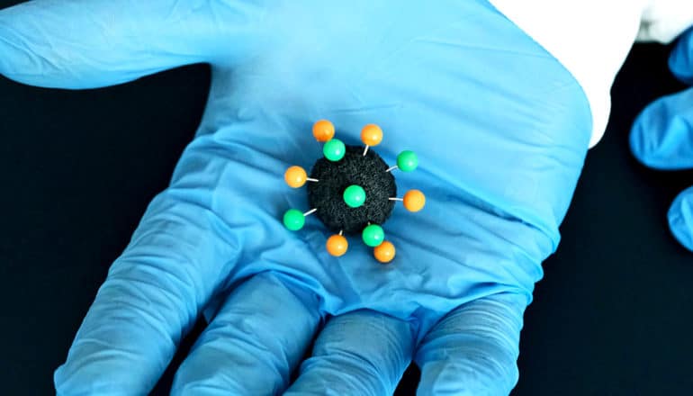 A small black, green, and orange model of the coronavirus sits in a blue-gloved hand