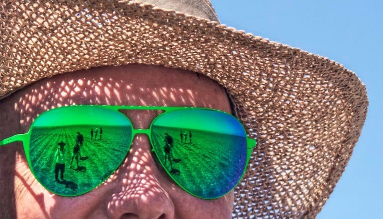 A person wears a woven straw hat and bright green sunglasses, their face red from heat. Workers on a farm field are visible in the glasses' reflection