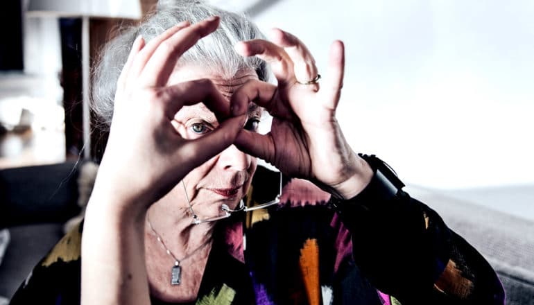 An older woman looks at the camera through her hands, shaped like glasses