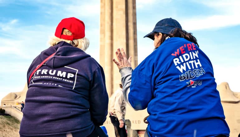 A Trump supporter and a Biden supporter in campaign clothing talk near a monument