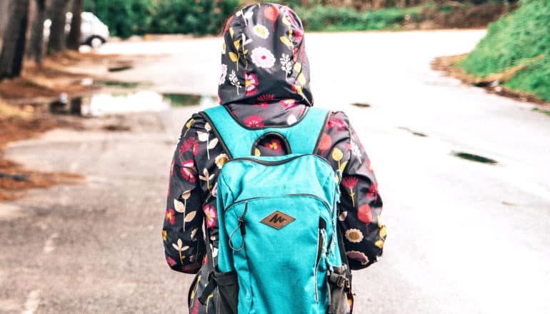 A young person in a raincoat with flowers on it walks to school wearing a blue backpack