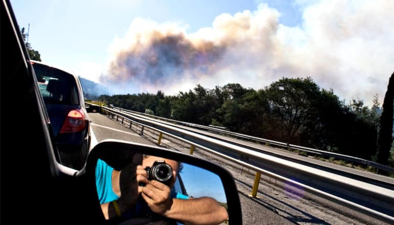 A man snaps a photo of smoke rising from a distant forest as he rides in a car, his camera visible in the rear-view mirror