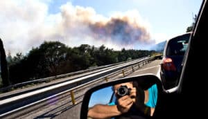 A man snaps a photo of smoke rising from a distant forest as he rides in a car, his camera visible in the rear-view mirror