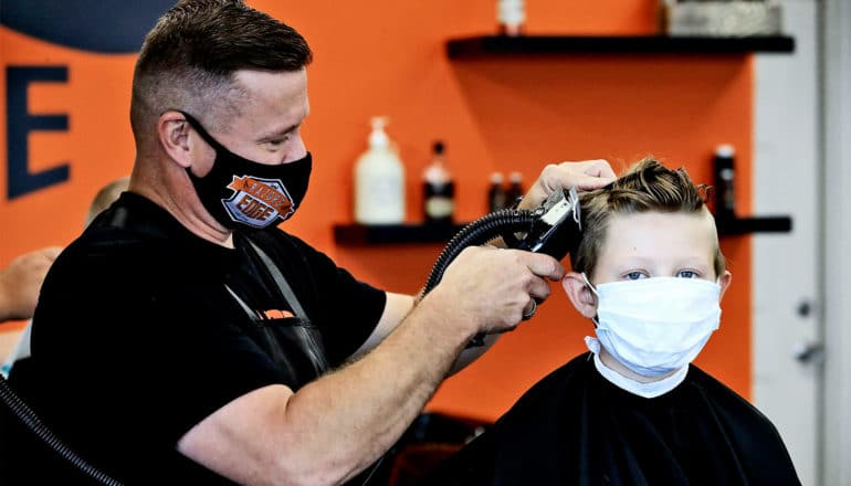 A man in a mask cuts a boy's hair as he sits in a barber's chair with a white mask on