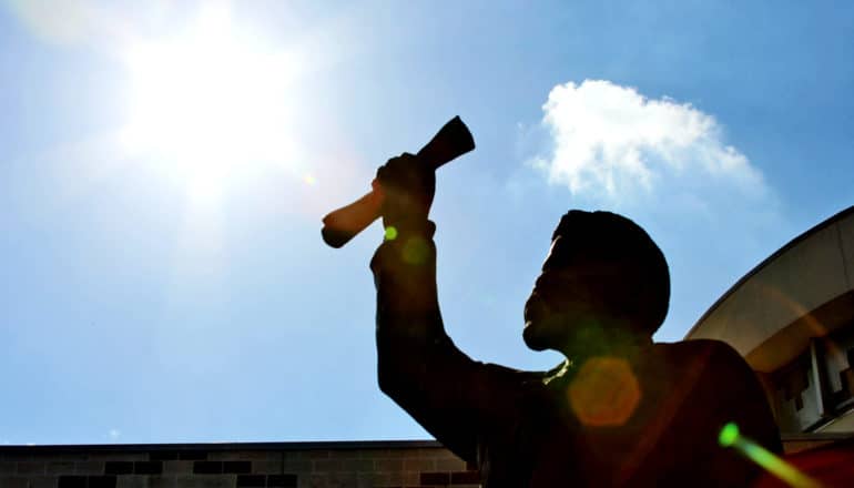 A statue in shadow holds up a rolled up document towards the sun