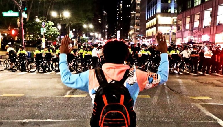 A young Black man stands with his hands up looking at a wall of police officers in front of him on the street