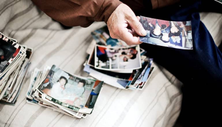 An old man sits on a bed looking at piles of old photos