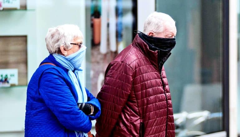 A woman in blue with a scarf around her face and a man in a red jacket with a black piece of cloth over his face walk down the sidewalk