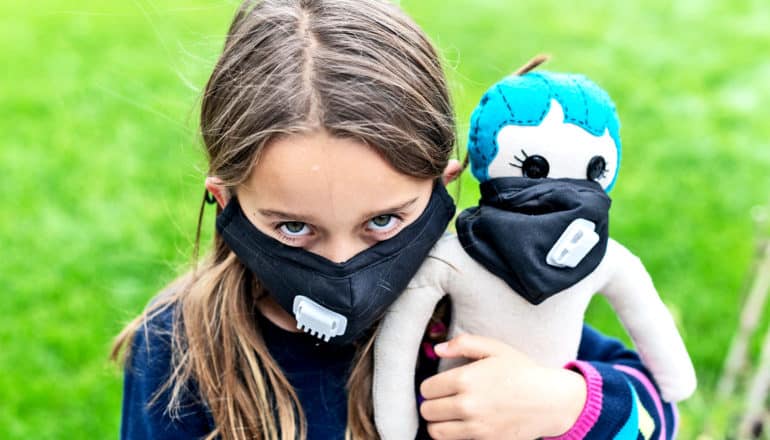 A young girl wears a black face mask while holding a doll with a similar black face mask