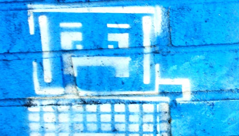 A computer with a frowning face is painted on a bright blue brick wall in white