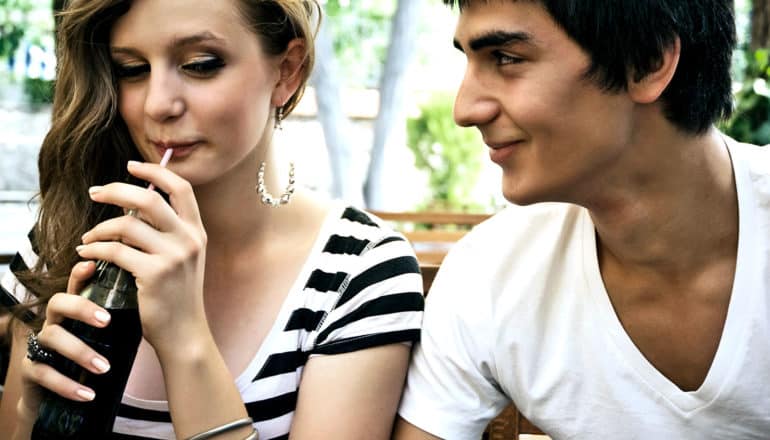 A young woman sips from a soda while looking sideways while smiling over at a man sitting next to her looking at her and smiling